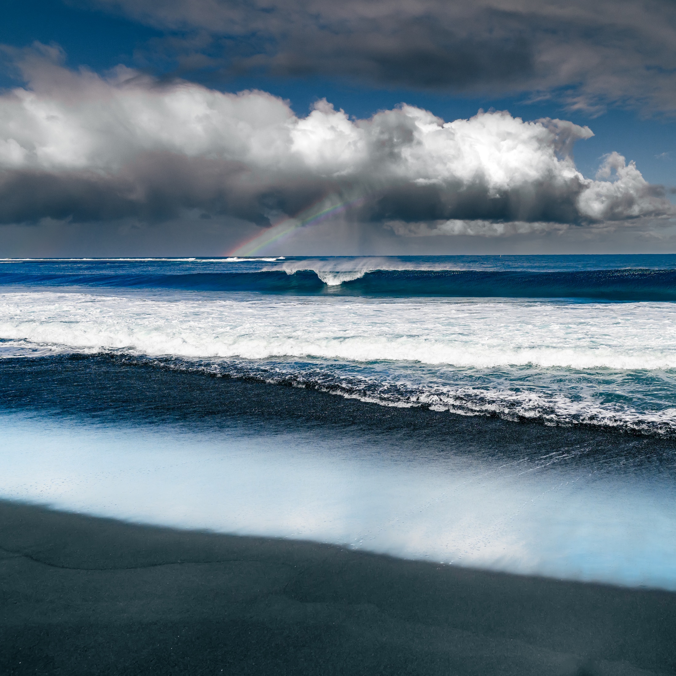 Ocean waves rolling toward a dark shoreline beneath storm clouds and a faint rainbow on Réunion Island