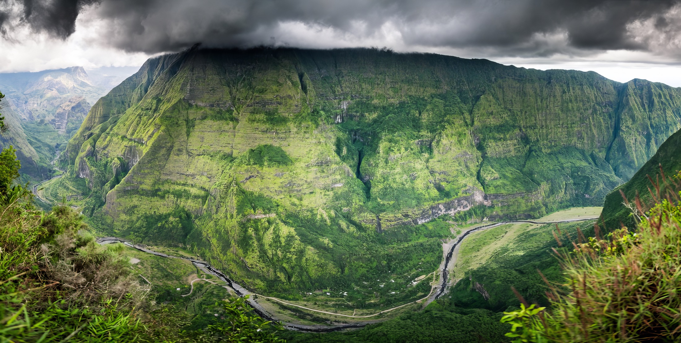 A vast steep-sided green valley beneath low dark cloud with roads winding across the valley floor on Réunion Island