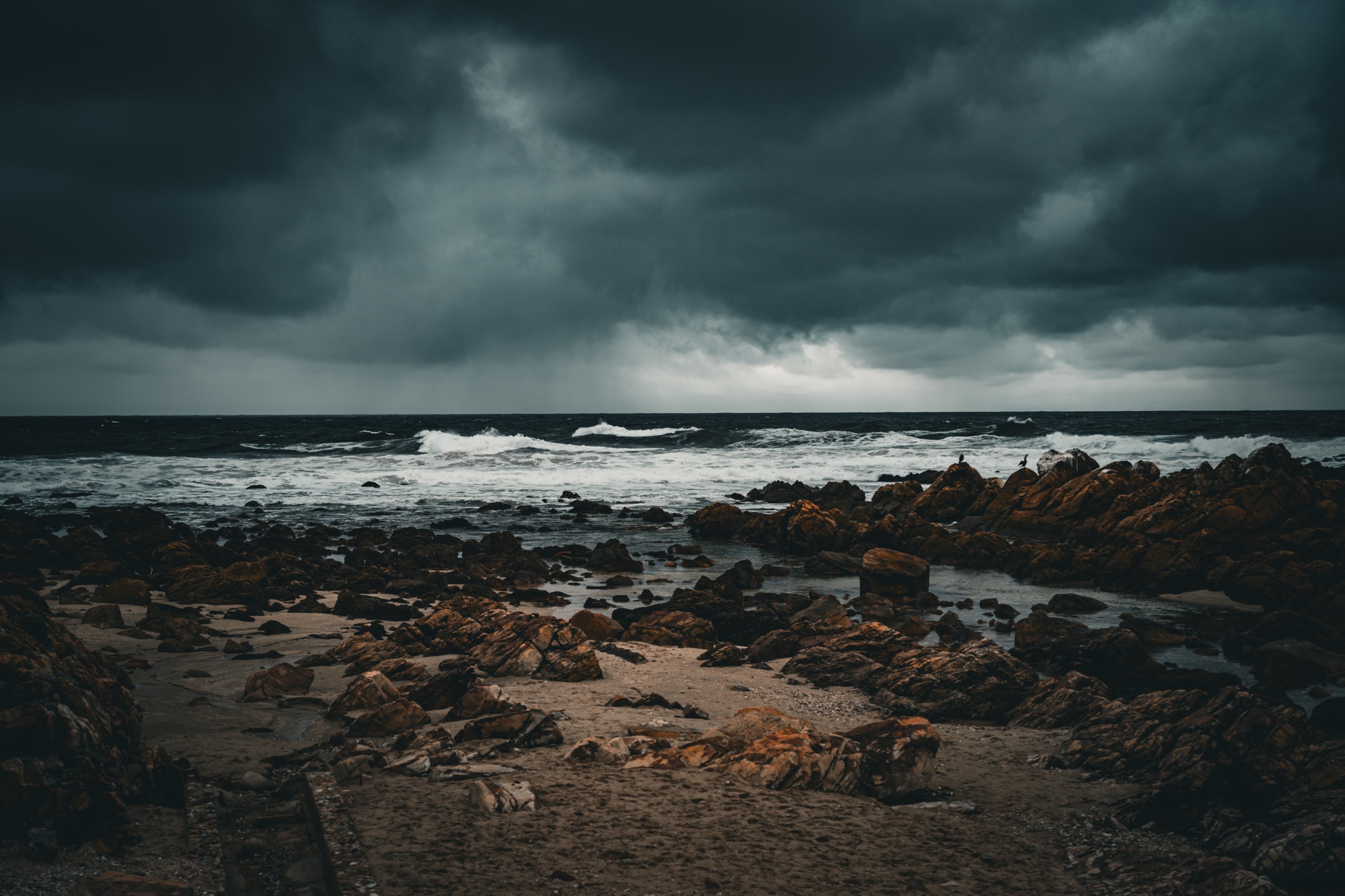 A rough grey ocean breaking over dark jagged rocks beneath a heavy stormy sky