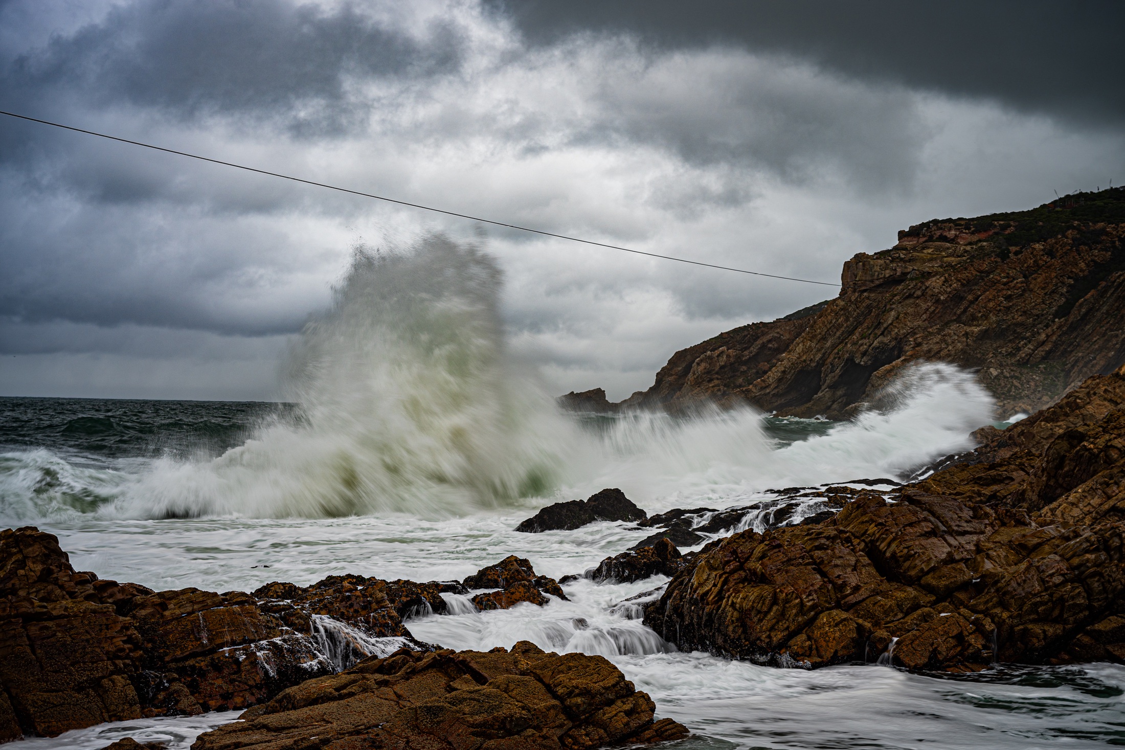 A large green-grey wave exploding into white spray between jagged rocks beneath dark coastal clouds