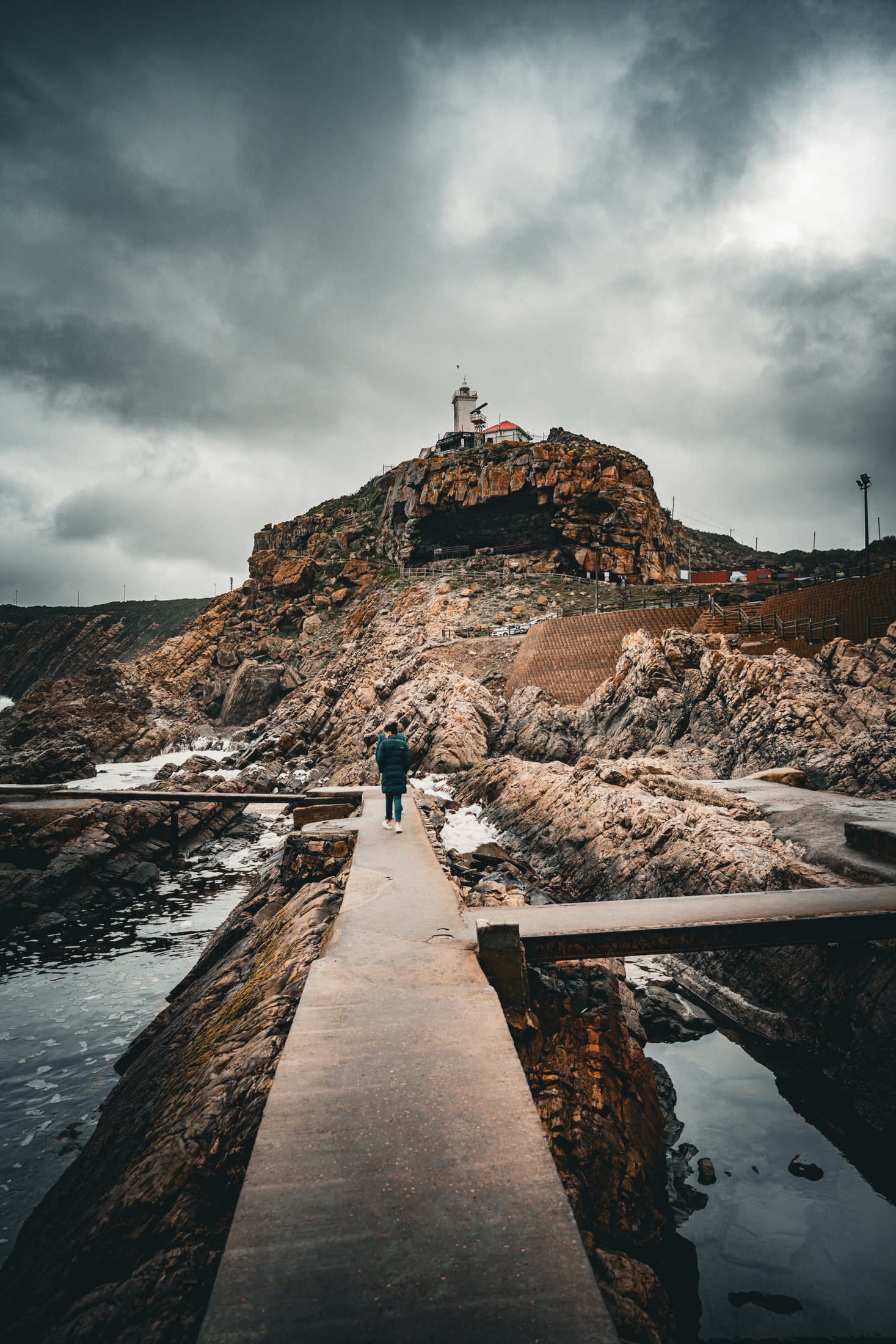A narrow concrete walkway crossing rocky tidal pools toward a lighthouse on the cliff at The Point in Mossel Bay