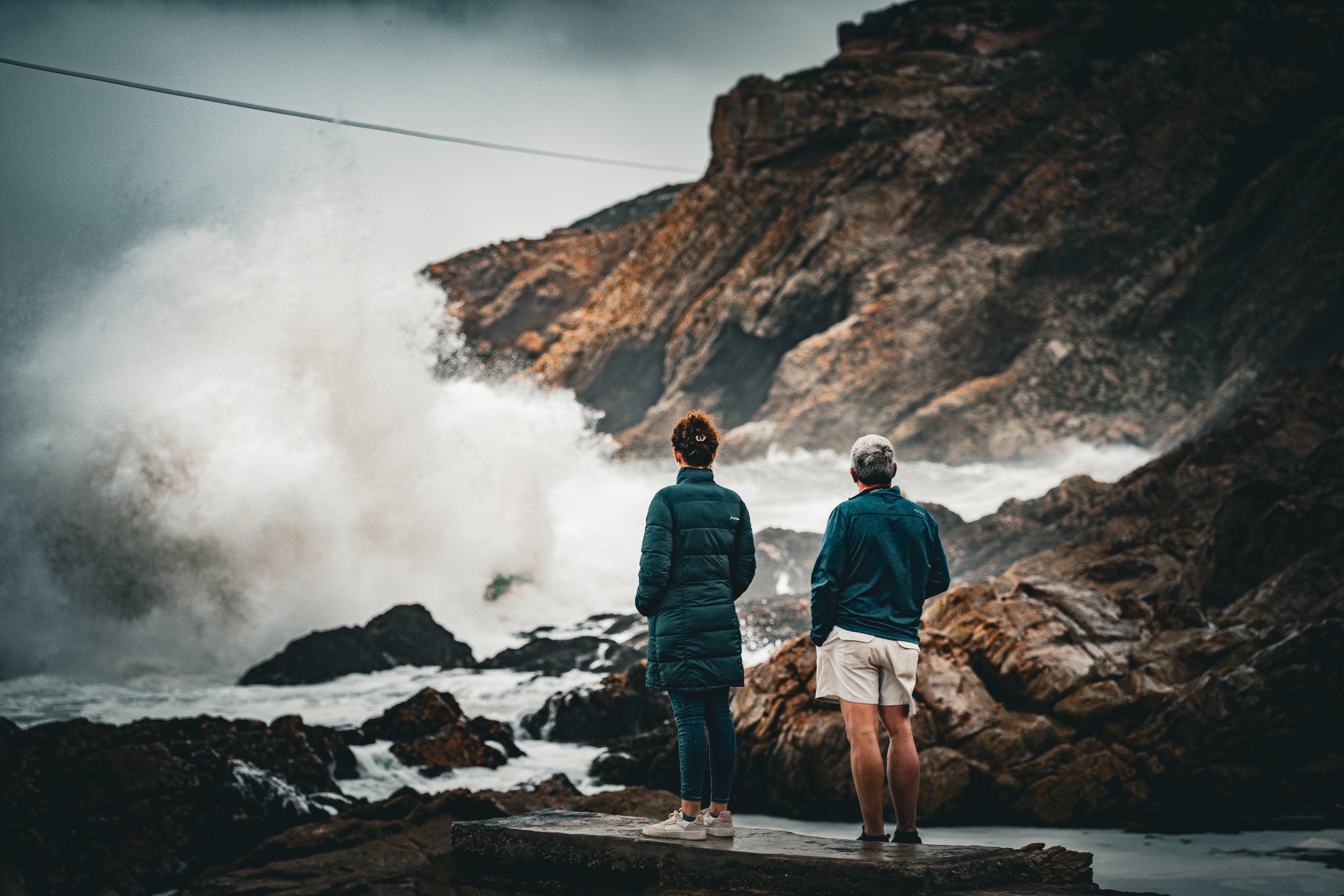 Two people standing on a concrete ledge as a large wave crashes against the rocks at The Point in Mossel Bay
