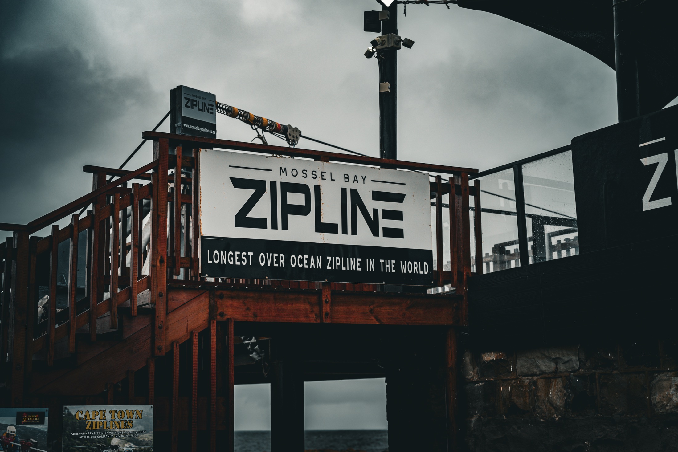 The Mossel Bay Zipline entrance sign above a wooden coastal structure beneath dark clouds