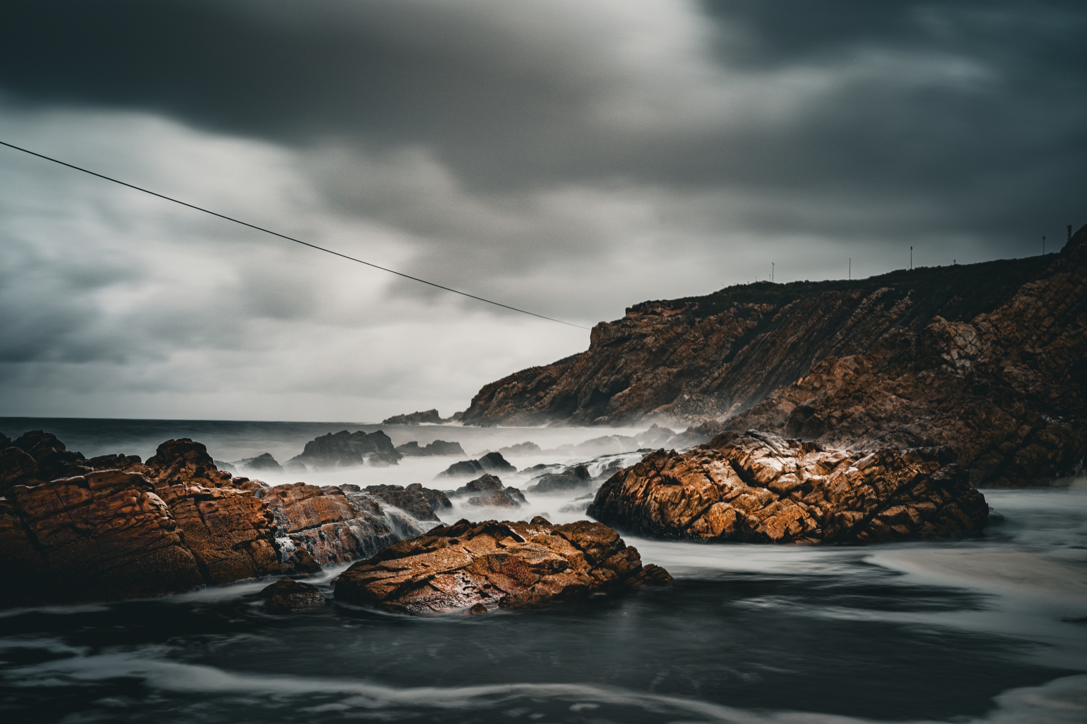 Dark seawater flowing between orange-brown coastal rocks beneath a moody overcast sky