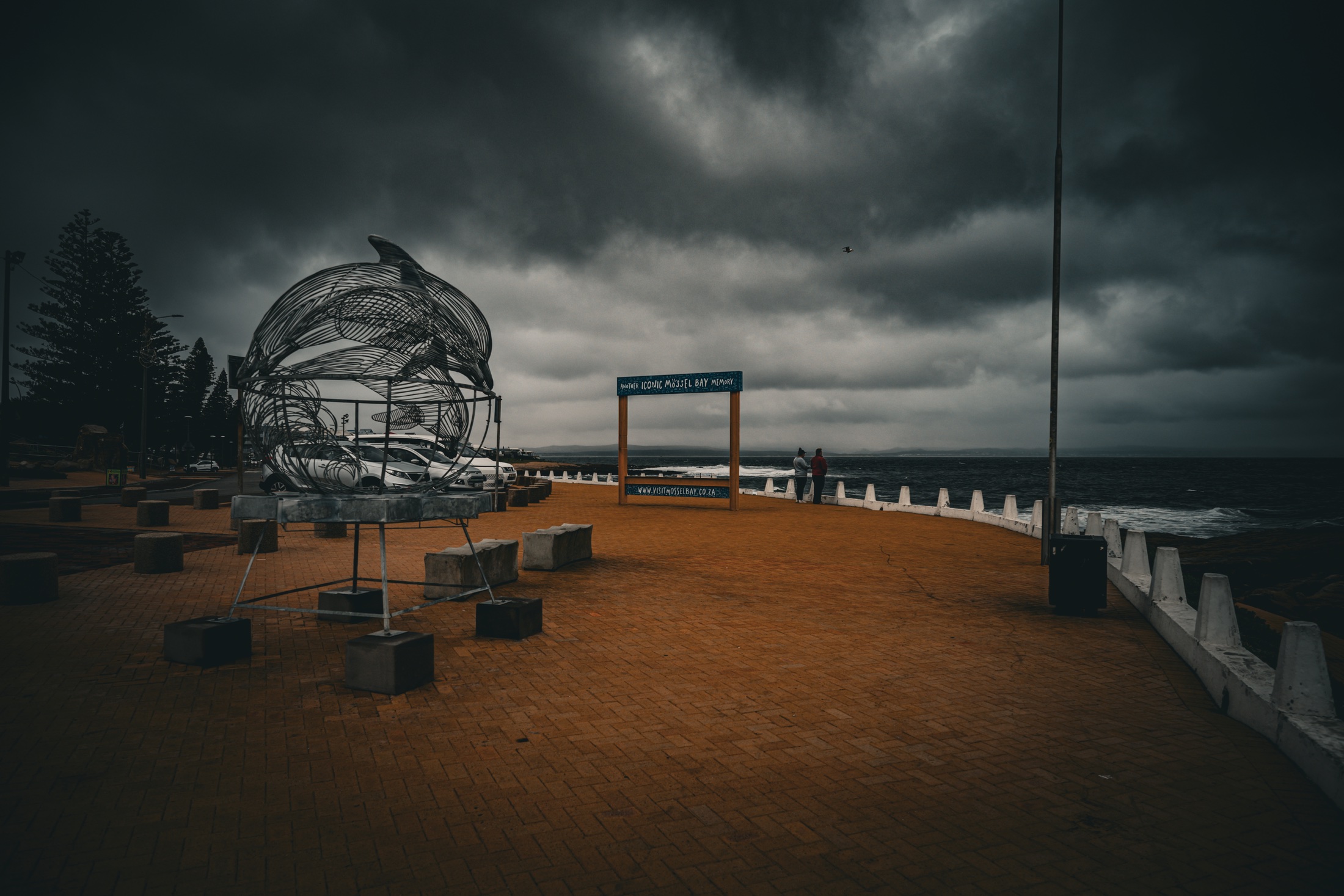 The paved promenade at The Point in Mossel Bay with a large metal dolphin sculpture, the ocean and dark storm clouds