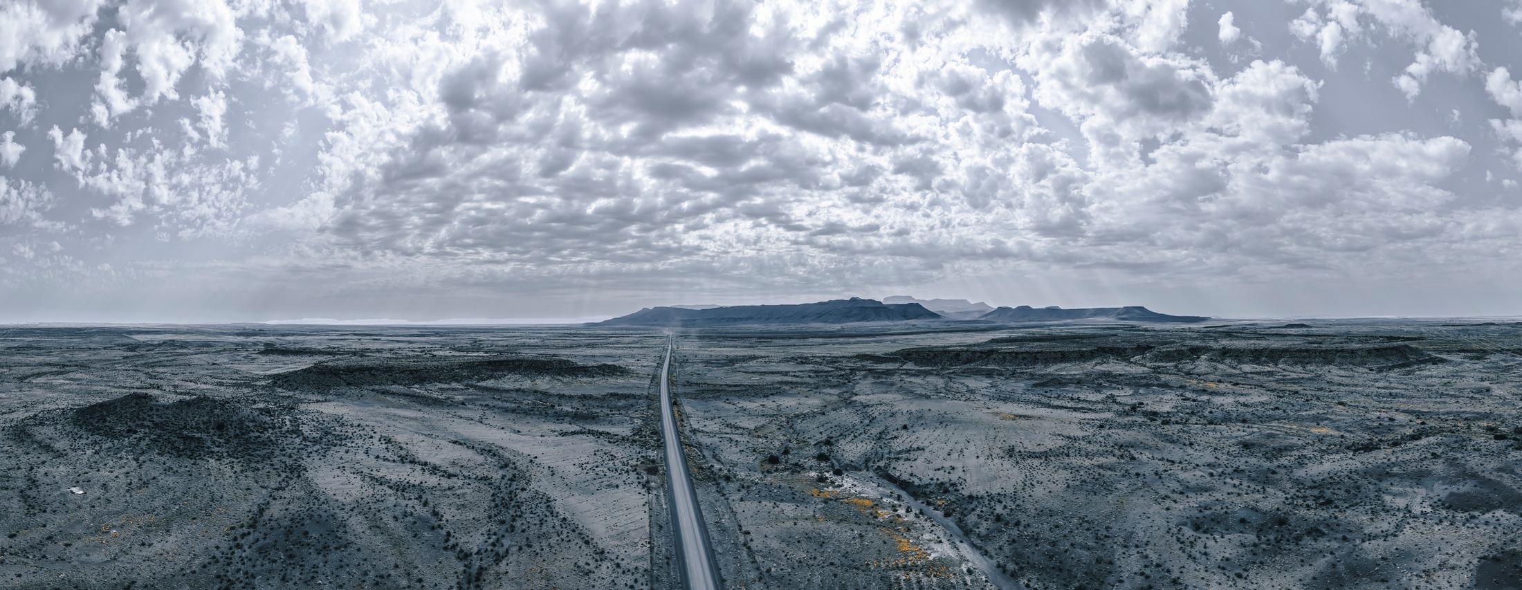 A black-and-white panorama of a straight road running through an expansive landscape beneath scattered clouds