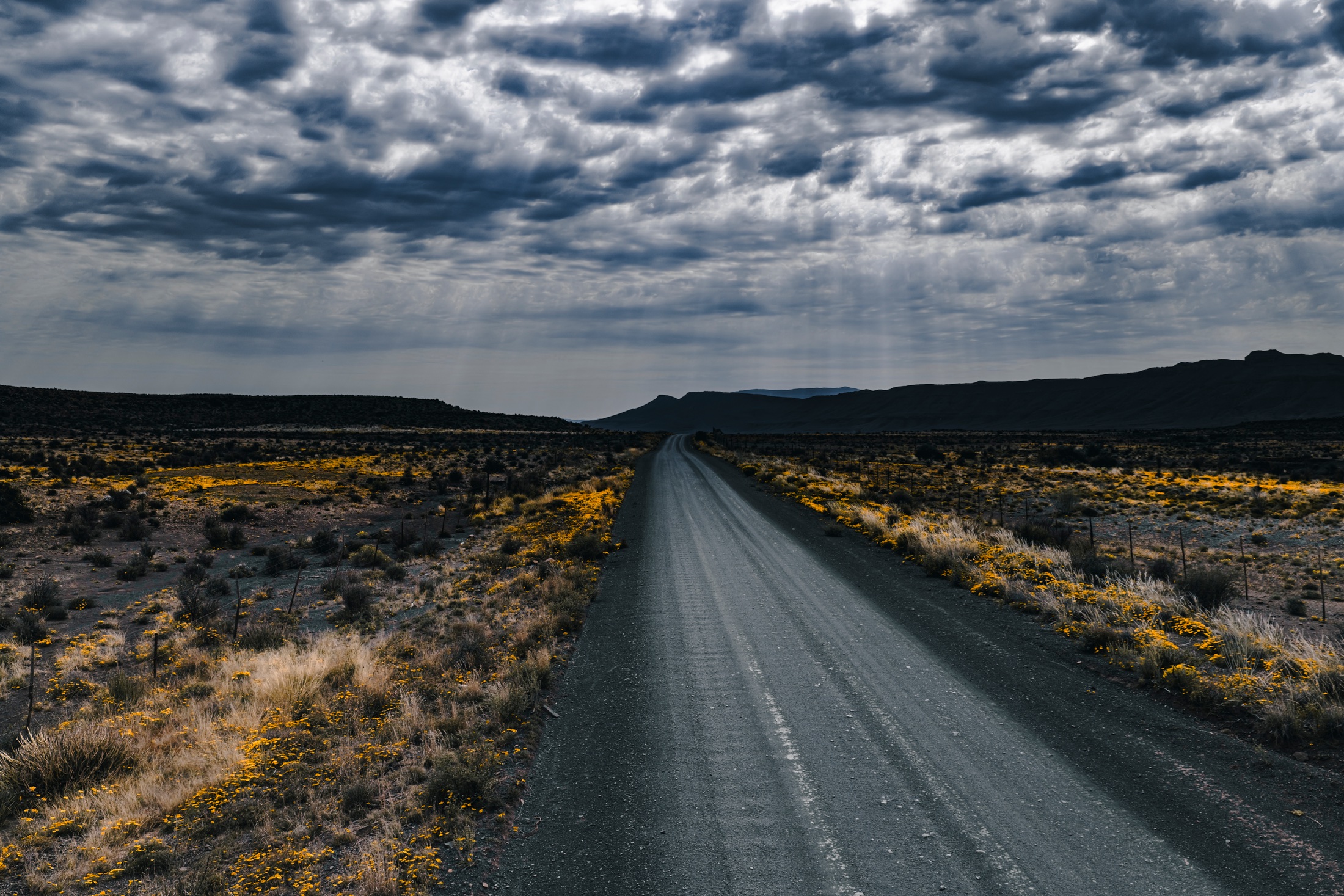 A straight gravel road leading towards distant flat-topped hills beneath textured clouds