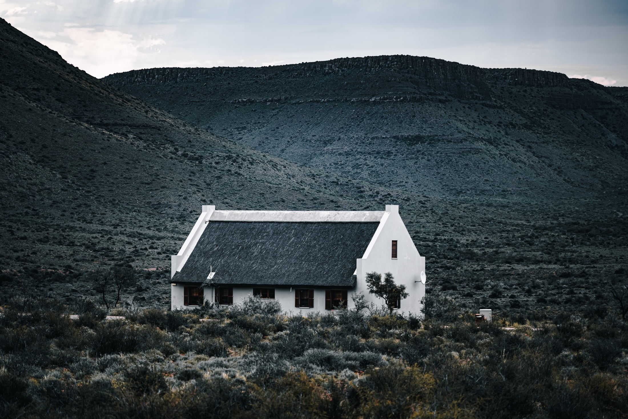 A small white cottage with a dark roof beneath a rugged, shadowed hillside